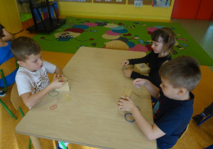Troje dzieci siedzi przy stole, w rękach trzymają gumki recepturki, które zakładają na kartonowe małe pudełko.